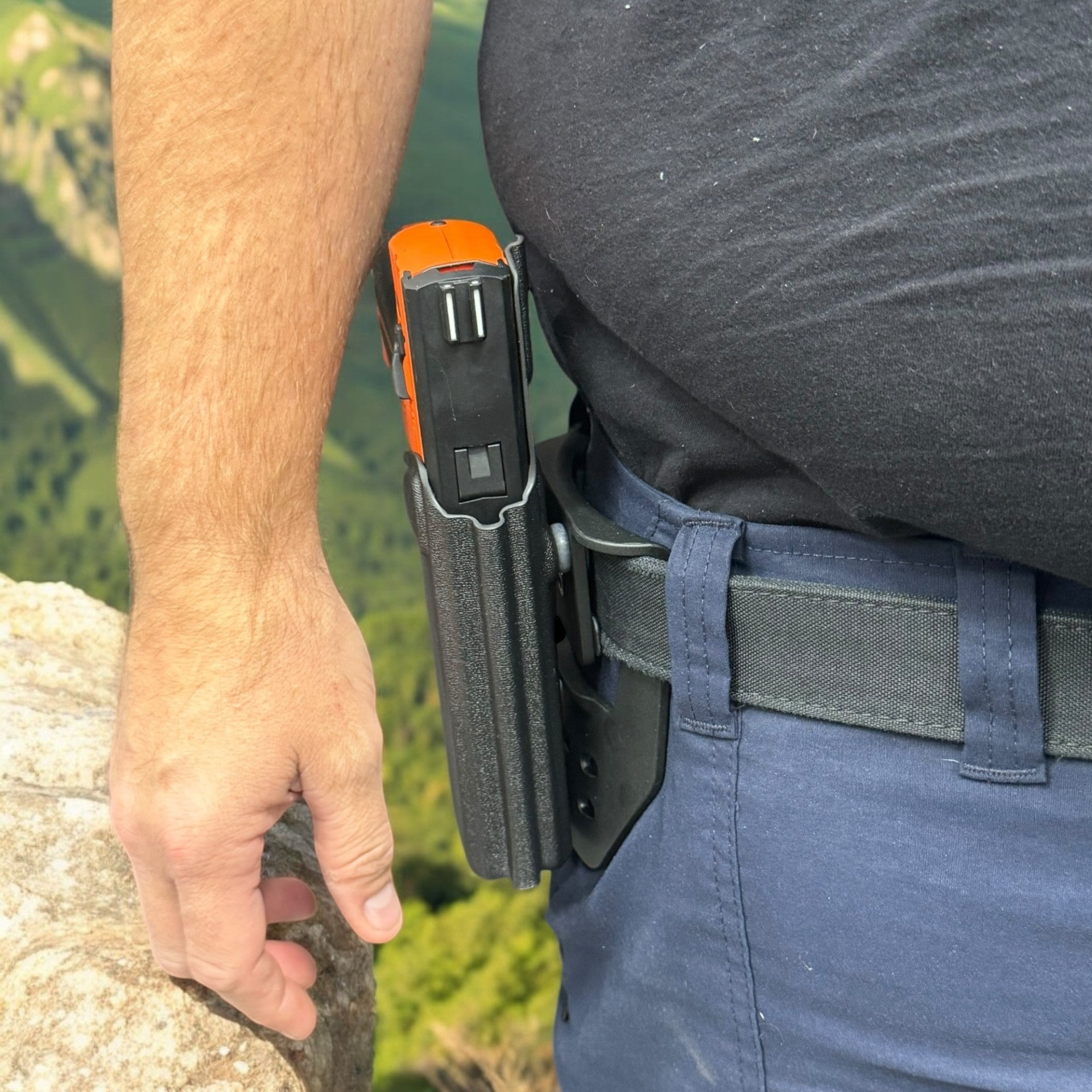 Person wearing a tactical belt with an orange and black device attached, outdoors.