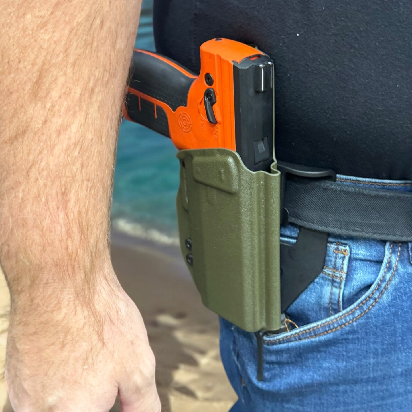 Orange and black gun in a green holster on a person's waistband with a blurred background.